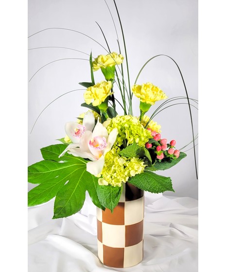 A modern style arrangement in a gold and ivory checkered vase. Green hydrangea, yellow carnations & white cymbidium orchid blooms accented with bear grass & hypericum berry.