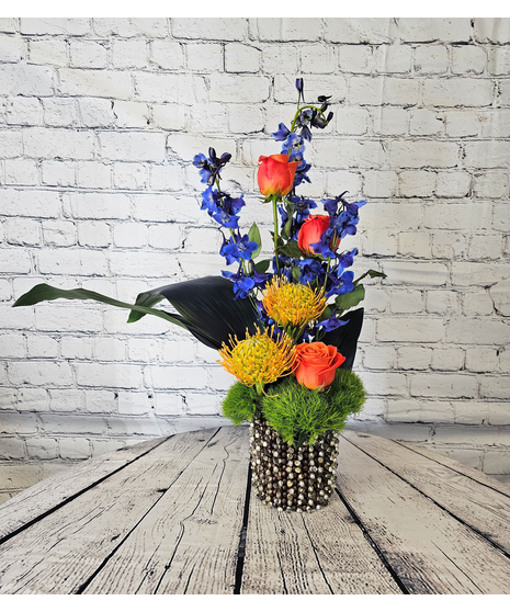 A modern container with orange roses, blue delphinium and pincushion protea. 